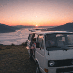 Furgoneta camper aparcada de frente, con la puerta trasera abierta, mirando hacia un espectacular amanecer sobre un mar de nubes en un valle gallego.